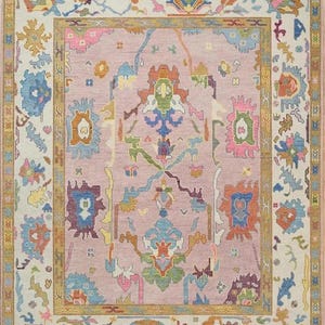 May include: A rectangular rug with a central field of muted pink, framed by a gold border and a cream-coloured outer border. The rug features intricate floral and geometric patterns in shades of blue, green, orange, and purple. The rug has a fringed edge.