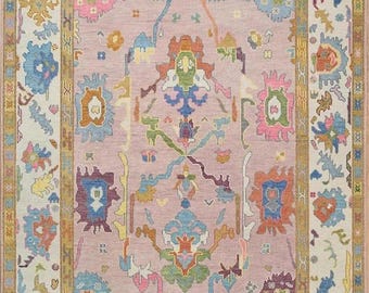 May include: A rectangular rug with a central field of muted pink, framed by a gold border and a cream-colored outer border. The rug features intricate floral and geometric patterns in shades of blue, green, orange, and purple. The rug has a fringed edge.