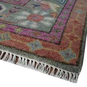 May include: Close-up of a rectangular rug with a detailed floral pattern. The rug features a gray-green background with pink, red, and blue floral designs. The border is a mix of pink and red, and the rug has white fringe.