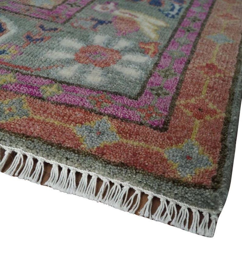 May include: Close-up of a rectangular rug with a detailed floral pattern. The rug features a gray-green background with pink, red, and blue floral designs. The border is a mix of pink and red, and the rug has white fringe.