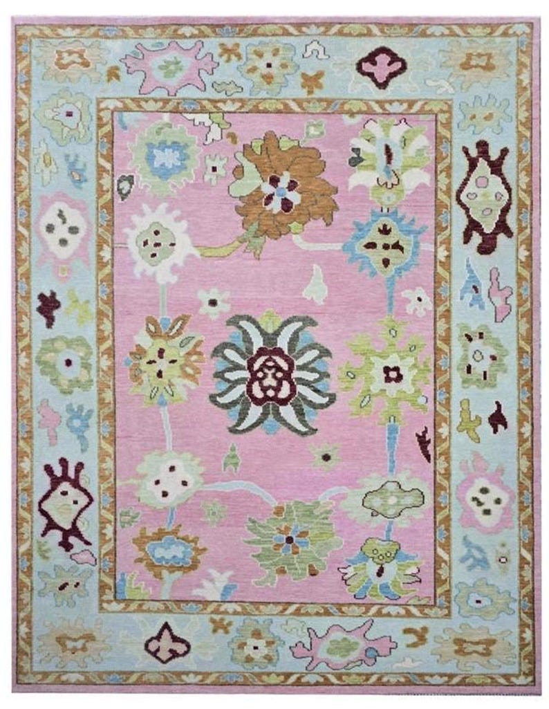 May include: A rectangular rug with a pink center and a light blue border. The rug features a floral pattern in shades of brown, green, and white. The border has a repeating design with floral and geometric elements. The rug's dimensions are not visible.