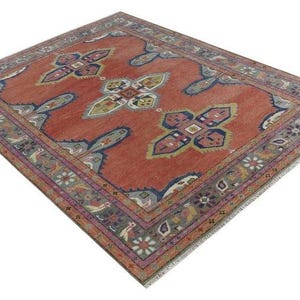 May include: A square, red rug with a detailed border and central design. The rug features a repeating pattern of stylized floral and geometric shapes in shades of blue, green, and cream. The border is a mix of floral and geometric elements.