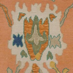 May include: Close-up of a peach-coloured rug with a detailed floral pattern. The design features a central motif in white, orange, and green, surrounded by blue, yellow, and pink floral accents. The rug has a textured appearance.