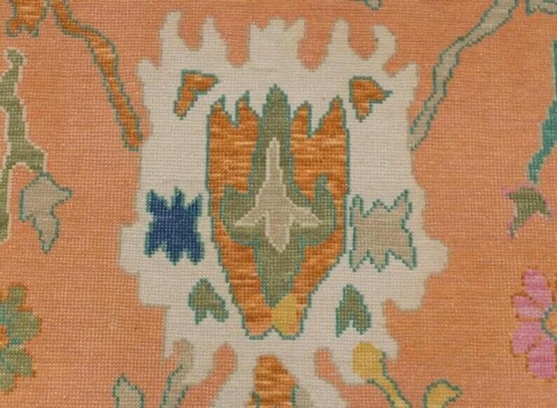 May include: Close-up of a peach-coloured rug with a detailed floral pattern. The design features a central motif in white, orange, and green, surrounded by blue, yellow, and pink floral accents. The rug has a textured appearance.