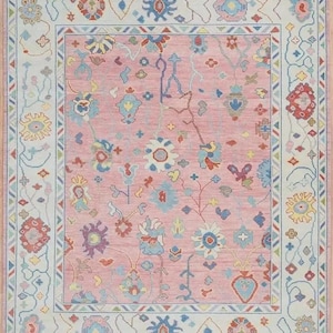 May include: A rectangular rug with a pink central field and a detailed border. The rug features floral and geometric patterns in shades of blue, red, yellow, and cream. The rug has a fringed edge.