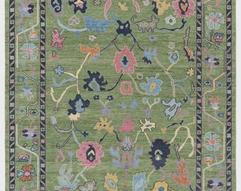 Op de afbeelding: Een rechthoekig, groen vloerkleed met een bloemenpatroon. Het vloerkleed heeft een centraal groen veld met kleurrijke, gestileerde bloemen in roze, blauw, geel en oranje. Een donkerblauwe rand omlijst het ontwerp en voegt contrast toe.