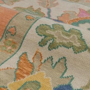 May include: Close-up of a rolled-up rug with a detailed floral pattern. The rug features a border in shades of green and beige, with a central design in orange, blue, and yellow. The rug's texture is visible, suggesting a woven construction.
