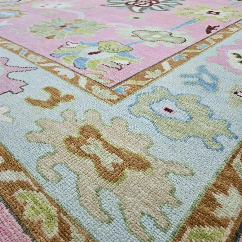 May include: Close-up of a decorative rug with a floral pattern. The rug features a light pink center with a light blue border. The design includes floral and geometric shapes in shades of green, brown, and beige.