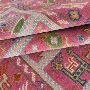 May include: Close-up of a folded pink rug with detailed geometric patterns. The rug displays a repeating design with shades of pink, blue, green, and orange. The rug's texture is visible, suggesting a soft, woven material.