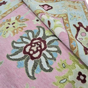 May include: A pink area rug with a floral pattern in shades of burgundy, blue, green, and tan. The rug has a border with a light blue background and floral designs. The rug is folded over, revealing the underside.