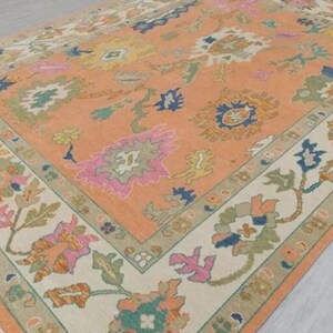 May include: A rectangular rug with a peach central field and a cream border. The rug displays a floral pattern in pink, blue, green, and yellow. The border has a repeating floral design. The rug has a fringe along the edge.