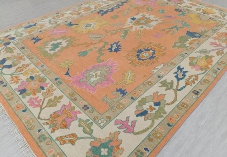 May include: A rectangular rug with a peach central field and a cream border. The rug displays a floral pattern in pink, blue, green, and yellow. The border has a repeating floral design. The rug has a fringe along the edge.