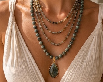 Puede incluir: Un collar de cuentas de varias hebras con tres capas internas de pequeñas cuentas de piedras preciosas translúcidas y multicolores, y una capa externa de cuentas redondas de color gris verdoso oscuro con un gran colgante en forma de lágrima facetada.