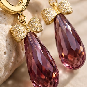 May include: A pair of gold hoop earrings featuring dangling teardrop-shaped mauve crystals. Each crystal is topped with a small gold bow encrusted with clear pavé stones. The earrings have a hinged closure mechanism at the top of the gold hoop.