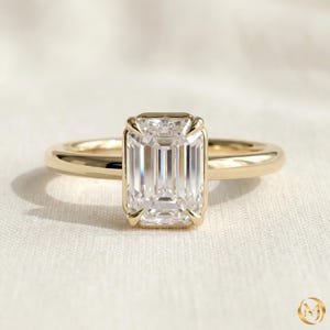 Op de afbeelding: Een gouden ring met een enkele, smaragdgeslepen heldere edelsteen in een zetting met vier pootjes. De ringband is glad en gepolijst en toont een klassiek ontwerp met een rechthoekige middensteen die het licht reflecteert op het gefacetteerde oppervlak.