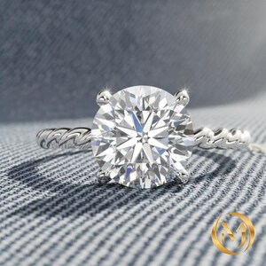 May include: A silver engagement ring with a large, round, clear gemstone. The band features a twisted rope design. The ring is set against a textured, light blue background.