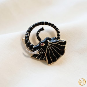 May include: A black and gold elephant brooch with a swirling trunk design. The brooch features a red eye and a tusk embellished with small, clear stones. The elephant's body is black enamel with gold trim.