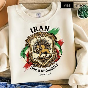 May include: Cream-colored sweatshirt featuring a detailed graphic of a golden lion holding a sword within a shield, with the word "IRAN" above and "SHIR O KHORSHEED" below. The design includes the Iranian flag colors and text in Farsi.