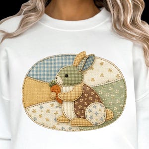 Puede incluir: Sudadera blanca con un diseño de conejo de patchwork. El conejo, sosteniendo una zanahoria, está sobre un fondo ovalado azul, verde y amarillo. El conejo está hecho de varias telas estampadas, creando una apariencia texturizada.