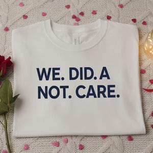 May include: A white t-shirt with the text "WE. DID. A NOT. CARE." in navy blue. The shirt is folded on a textured, light-colored surface, with a red rose, candles, and decorative lights.