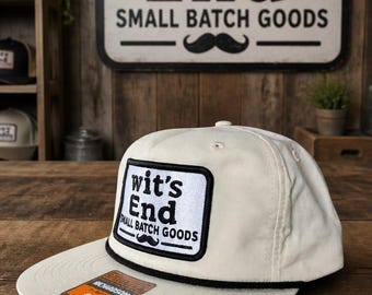 May include: An off-white baseball cap with a black rope detail across the brim. A rectangular white patch on the front features the black text "wit's End SMALL BATCH GOODS" and a black mustache icon. A small orange sticker is on the brim.