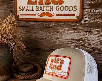 May include: A beige and white trucker hat featuring a rectangular patch with the orange and brown text "wit's End SMALL BATCH GOODS" and an orange mustache icon. The hat has a black sticker on the brim with the text "112 The Original MD 20".