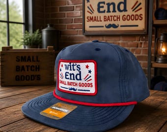May include: A navy blue baseball cap with a flat brim and a red rope detail across the base of the crown. A rectangular patch on the front features the text wit's End SMALL BATCH GOODS in blue and red, accented with stars and a blue mustache.