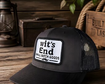 May include: A charcoal gray and black trucker hat featuring a rectangular white patch on the front. The patch displays the black text "wit's End" above the words "SMALL BATCH GOODS" and a small mustache icon centered between two horizontal lines.