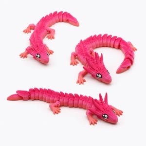 May include: Three articulated dragon figurines in a vibrant hot pink color. Each creature features a segmented body, four small legs, and a detailed head with large, expressive black and white eyes. The figures are posed in different positions.