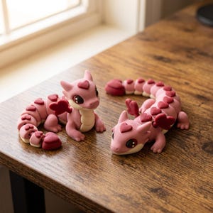 May include: Two articulated dragon figurines in shades of light pink and dark red. Each dragon features a heart-shaped crest on its head, heart-shaped wings, and a segmented tail with heart-shaped scales along its back and a cream-colored belly.
