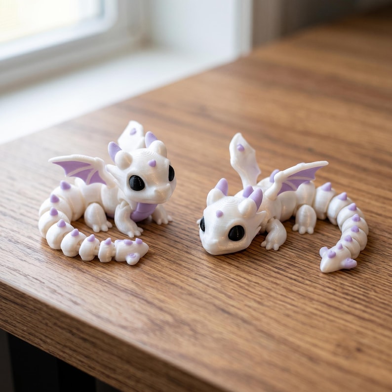 May include: Two articulated dragon figurines featuring white bodies with light purple accents on the wings, horns, and tail spikes. Each dragon has large black eyes, a segmented tail, and small wings, crafted with a smooth finish and visible joints.