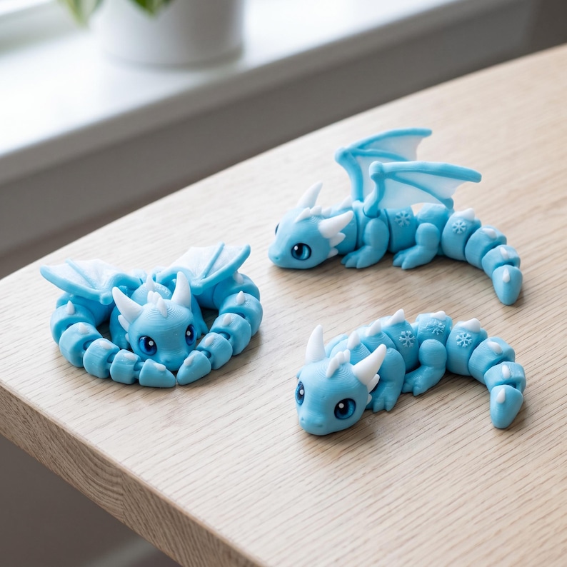 May include: Three articulated dragon figurines in light blue with white accents. One dragon is curled into a circle, while the other two are in crawling positions. Each dragon features small white snowflake patterns on its back and tail segments.