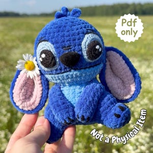 Könnte beinhalten: Handgefertigte blaue Stitch-Häkelpuppe mit großen schwarz-weißen Augen, einer schwarzen Nase und rosa gefütterten Ohren. Eine weiße Gänseblümchen steckt hinter einem Ohr. Der Text "Pdf only" und "Not a Physical Item" ist enthalten.