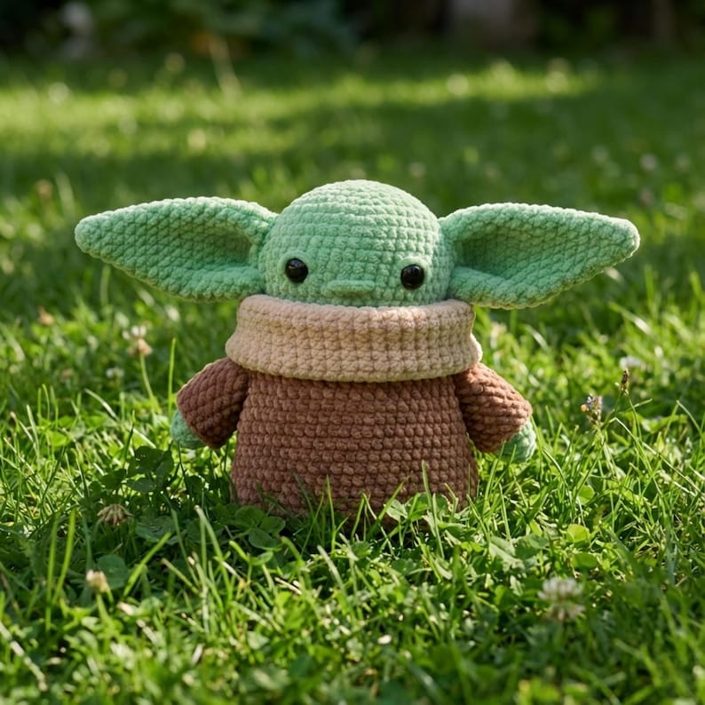 Op de afbeelding: Een gehaakte pop van een personage met grote oren, groene huid en een bruin gewaad. De pop zit in een bed van groen gras. De pop heeft zwarte ogen en een bruine kraag.