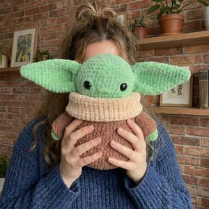Op de afbeelding: Een handgemaakt gehaakt pluche speelgoed van een personage met grote oren, groene huid en een bruine jurk. Het speelgoed heeft zwarte ogen en een beige kraag. Het pluche wordt vastgehouden voor een persoon die een blauwe trui draagt.