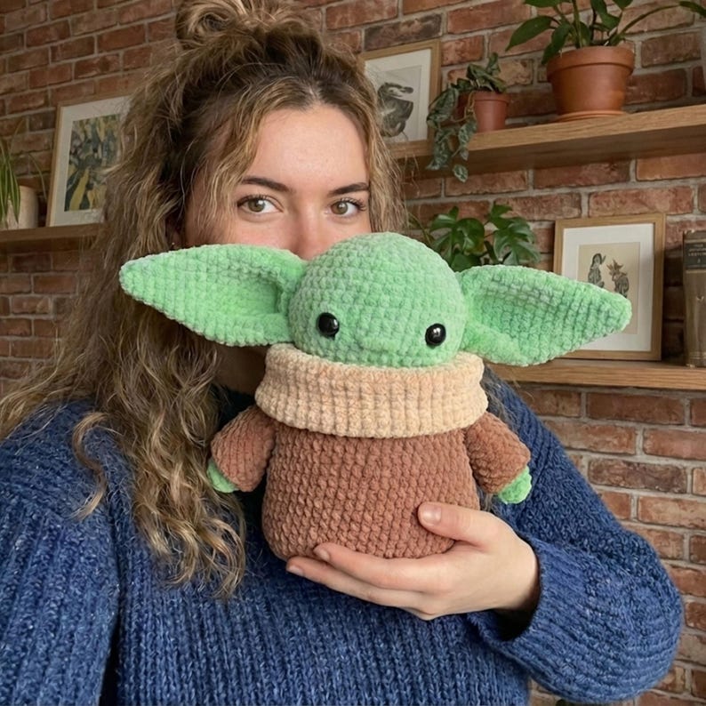 Op de afbeelding: Een handgemaakte groene en bruine gehaakte pop met grote oren, zwarte ogen en een beige kraag. De pop wordt vastgehouden voor een persoon die een blauwe trui draagt. De pop is een personage uit een populaire sciencefictionfranchise.