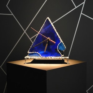 Könnte beinhalten: Eine dreieckige Uhr aus Buntglas in tiefem Blau. Sie hat silberfarbene Verzierungen und zarte Beine. Die Zeiger sind kupferfarben. Die Uhr steht auf einem schwarzen Podest vor einem geometrischen Hintergrund.