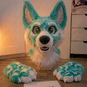 Peut inclure: Un loup en fourrure turquoise et blanche, des pattes et un panneau avec l'inscription "KRAUS". La tête de loup a de grands yeux verts et un nez noir. Les pattes ont un motif turquoise et blanc.