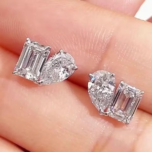 May include: A pair of sparkling diamond earrings. Each earring features a pear-shaped diamond paired with an emerald-cut diamond. The diamonds are set in a silver or white gold setting, reflecting light.