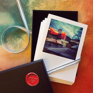 May include: A stack of white cards featuring a city scene, tied with a dotted ribbon, rests in a black box. Another black box with a red RSVP London seal is in the foreground. The background is a mix of orange, green, and yellow.