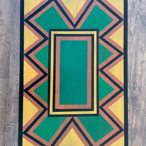 Puede incluir: Una mesa rectangular de madera con un diseño geométrico. El diseño presenta un rectángulo verde central enmarcado por bordes marrones y negros. El patrón exterior incluye zigzags amarillos, verdes y marrones, todos delineados en negro. Los bordes de la mesa son negros.