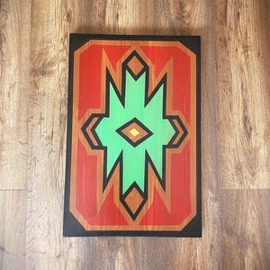 May include: Geometric abstract painting on a wooden panel. The artwork features a central green shape with a yellow center, surrounded by red and orange geometric elements, all framed in black.