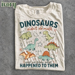 May include: An off-white t-shirt featuring a graphic of five colorful dinosaurs, mathematical equations, and the text "DINOSAURS didn't do math and we know what HAPPENED TO THEM". The design includes a triceratops, T-Rex, and other prehistoric creatures.