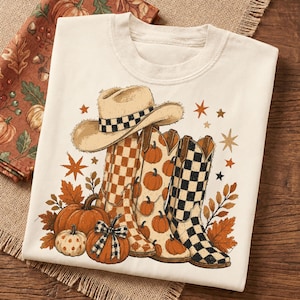 Könnte beinhalten: Ein cremefarbenes T-Shirt mit einem Grafikdruck, der einen beigen Cowboyhut, drei Westernstiefel mit Karomustern und Kürbismotiven sowie mehrere orangefarbene Kürbisse zeigt. Das Design ist mit braunen Herbstblättern und kleinen Sternen verziert.
