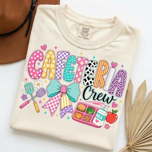 Può includere: Una t-shirt color crema con la scritta CAFETERIA Crew in lettere a bolle colorate e decorate. Il design include un grande fiocco verde acqua, due spatole, un vassoio da pranzo rosa con cibo, un cartone di latte e una mela rossa.