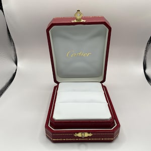 以下が含まれることがあります： 開いた赤い革製のジュエリーボックス。金色の王冠クラスプが付いています。内側は白で、金色の「Cartier」のロゴが入っています。リングやその他の小さなジュエリーを収納するように設計されています。