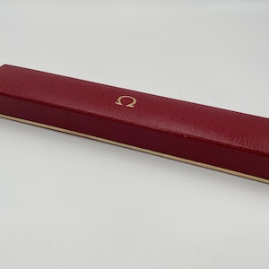 May include: A rectangular, claret-coloured box with a gold-coloured trim. The box has a textured surface and a gold-coloured Omega logo on the top. Likely for a watch or jewellery.