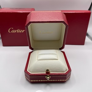 Puede incluir: Caja de joyería de cuero rojo abierta con interior blanco. La caja es octogonal y tiene detalles dorados. El logotipo de Cartier es visible en la caja y la tapa. Una segunda caja es visible en el fondo.
