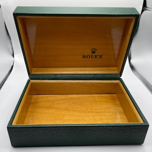 Könnte beinhalten: Eine offene, rechteckige, dunkelgrüne Box mit Holzinnenraum. Auf der Innenseite des Deckels steht in schwarzer Schrift „ROLEX“ mit einem Kronensymbol darüber. Die Box ist leer.