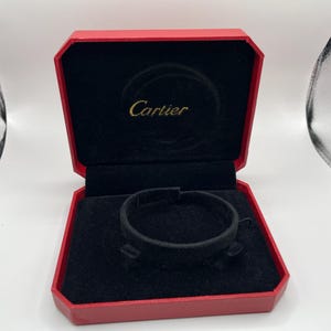 Puede incluir: Caja Cartier roja abierta con interior negro y pulsera negra. El logotipo de Cartier está en letras doradas en el interior de la tapa. La caja está abierta, revelando la pulsera y el interior.